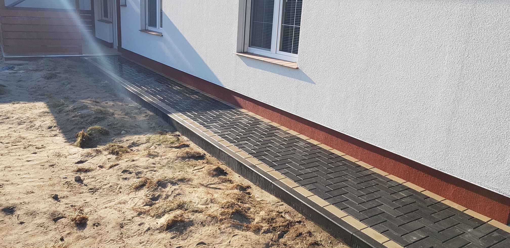 Ścieżka z kostki brukowej ułożona w jodełkę, z obrzeżami w kolorze piaskowym, prowadząca wzdłuż białej ściany budynku z oknami, na tle piaszczystego terenu.