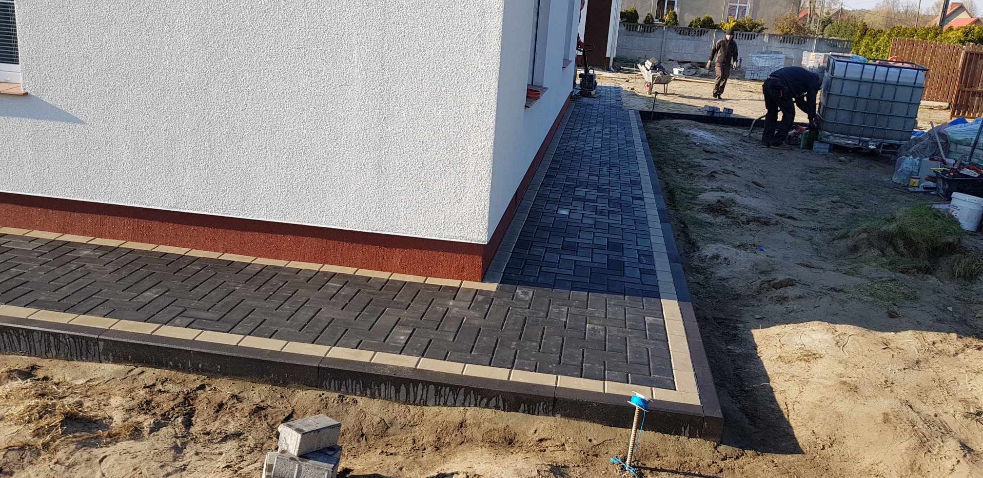 Ułożona ścieżka z kostki brukowej w odcieniach szarości i beżu, obramowana ciemniejszym obrzeżem, przylegająca do ściany budynku z widocznymi pracownikami w tle.