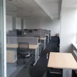 Długi korytarz biurowy z rzędami stanowisk pracy oddzielonych przegrodami, widoczne biurka z jasnymi blatami i szare krzesła obrotowe na ciemnej wykładzinie dywanowej, oświetlenie sufitowe i duże...