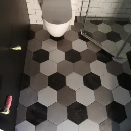 Wykończona łazienka z heksagonalną, szaro-biało-czarną mozaiką na podłodze, wisząca toaleta i fragment kabiny prysznicowej z widocznymi narzędziami glazurniczymi.