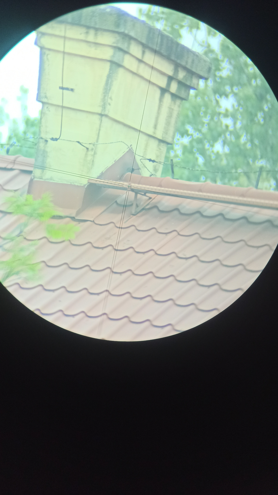 Zdjęcie dachu z kominem widziane przez lunetę geodezyjną, z widocznymi liniami celowniczymi i roślinnością w tle.