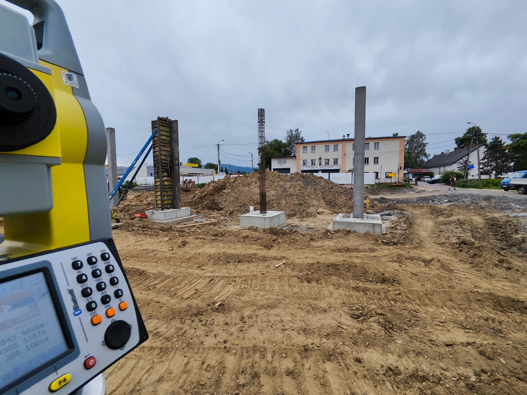 Prace ziemne pod budowę z widocznym betonowym fundamentem i wystającymi słupami zbrojeniowymi, w tle budynek mieszkalny oraz żółto-szary teodolit z klawiaturą na pierwszym planie.