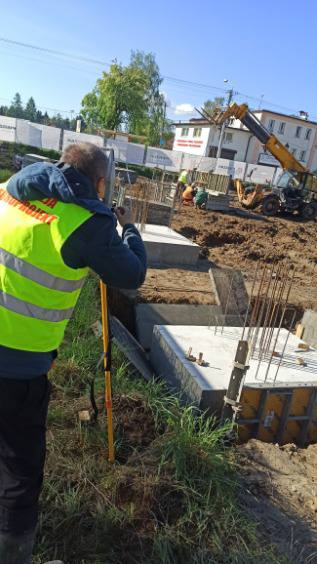 Prace geodezyjne na budowie: mężczyzna w żółtej kamizelce mierzy poziom fundamentów, w tle widoczny dźwig i ekipa budowlana.