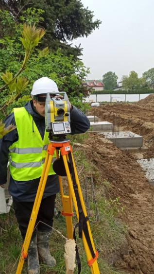 Pracownik w kasku i kamizelce odblaskowej obsługuje niwelator na budowie, w tle widoczne wykopane fundamenty.