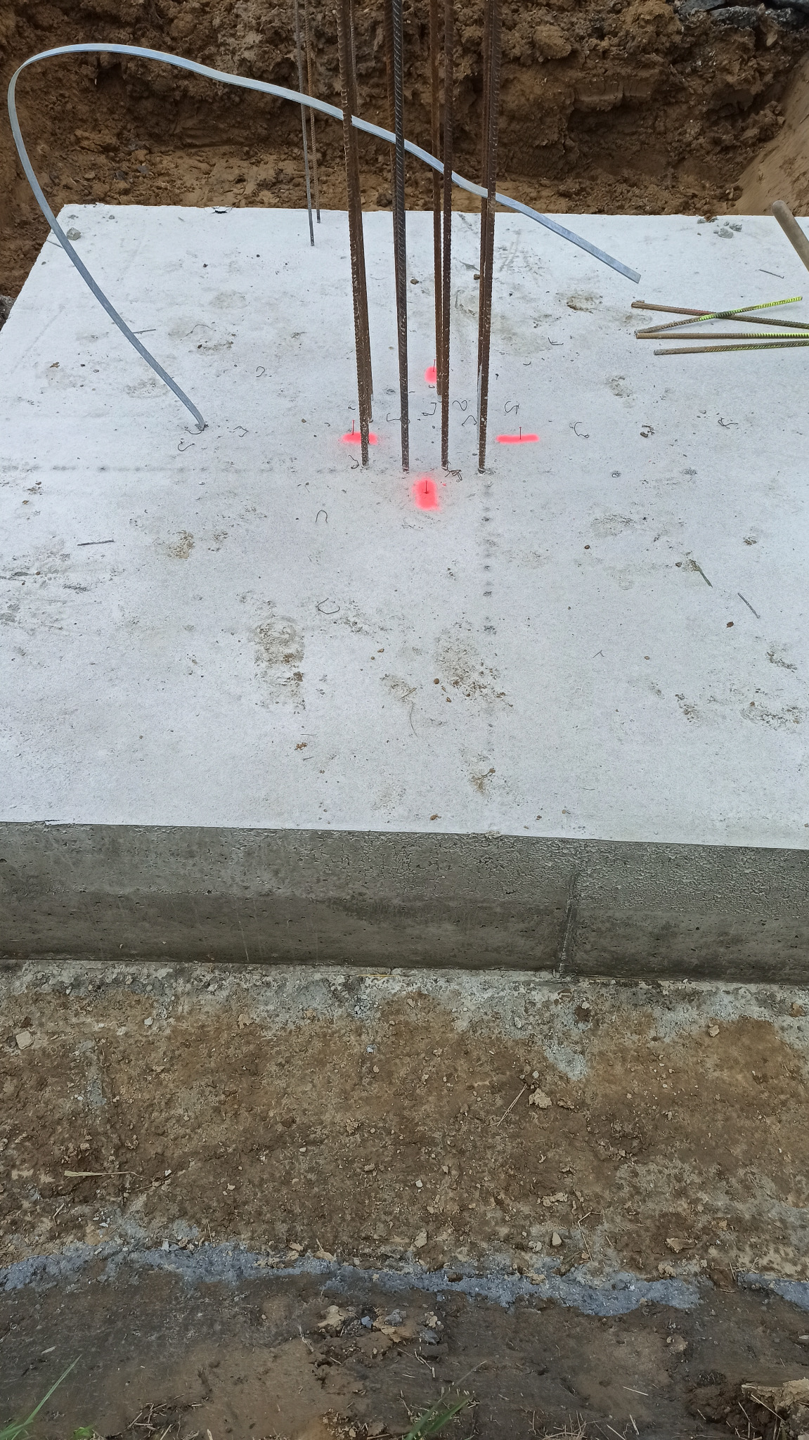 Betonowa płyta fundamentowa z wystającymi prętami zbrojeniowymi, oznaczona czerwonymi punktami pomiarowymi, w wykopie ziemnym.