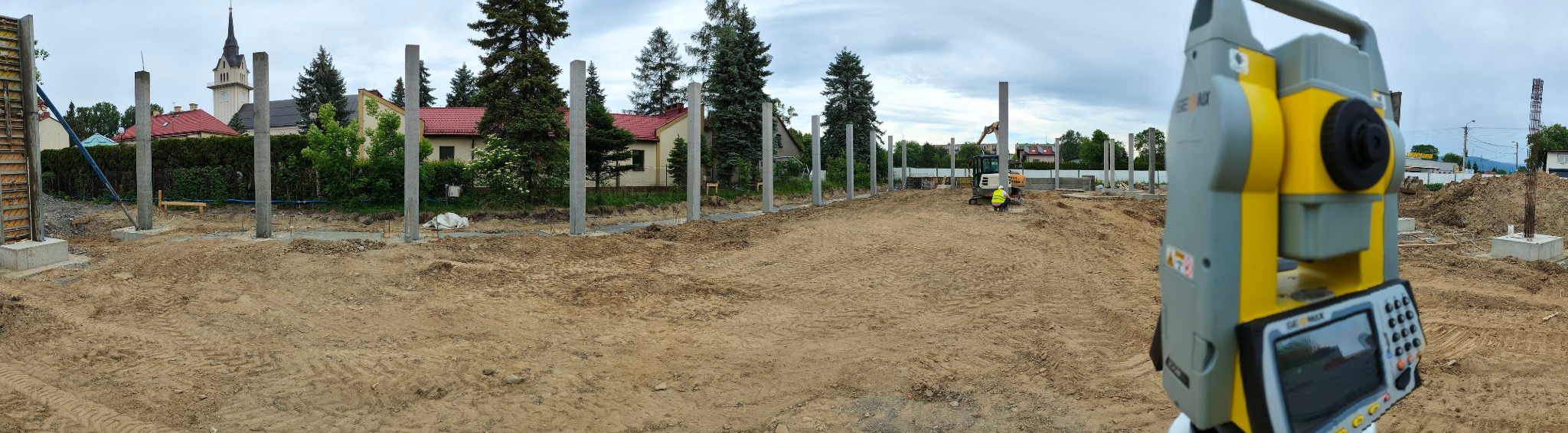 Budowa domu jednorodzinnego: widok na plac budowy z widocznymi betonowymi słupami, w tle kościół, a na pierwszym planie teodolit w kolorze żółto-szarym.