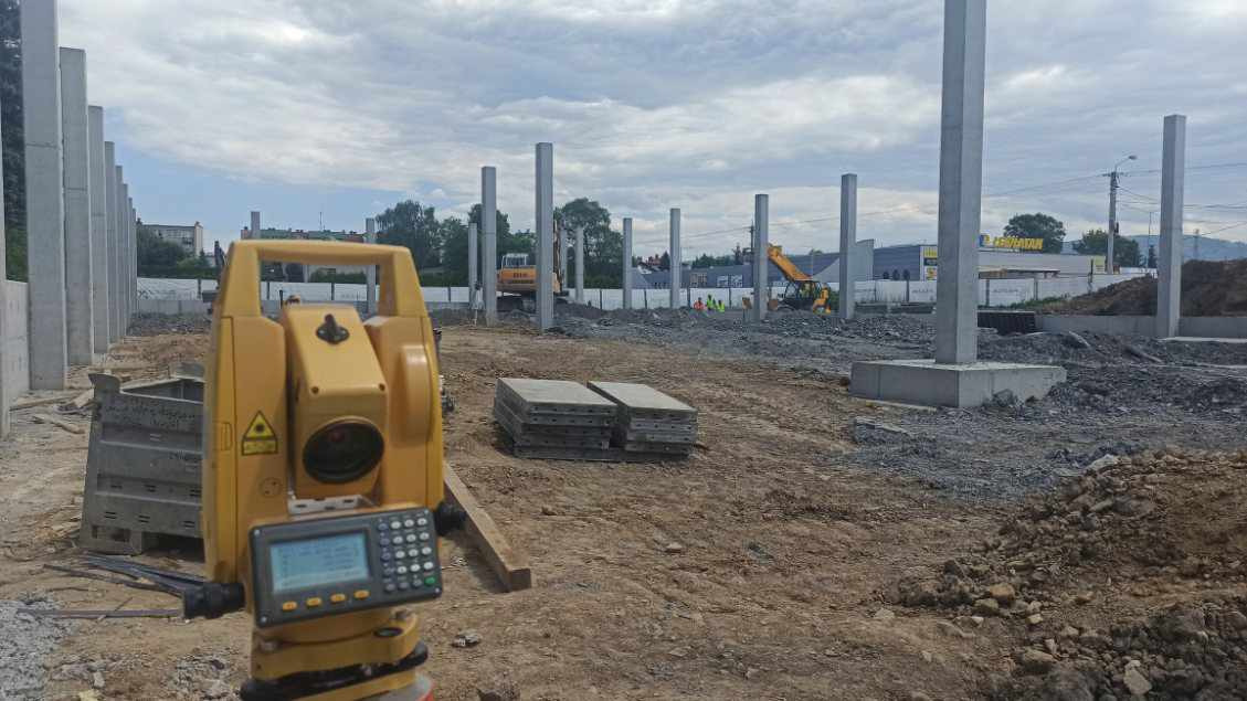 Żółty teodolit na placu budowy z widocznymi betonowymi słupami i maszynami budowlanymi w tle, pochmurne niebo.