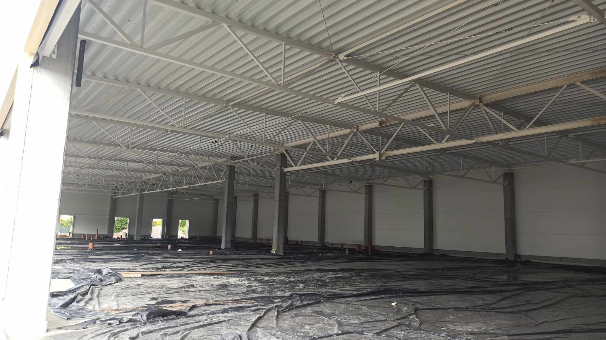 Wnętrze hali produkcyjnej w trakcie budowy, widoczne stalowe elementy konstrukcyjne dachu, betonowe słupy, folia budowlana na podłożu i przygotowane rury instalacyjne.