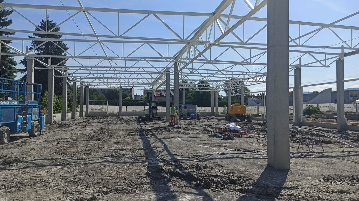 Wnętrze budowanego obiektu z widoczną stalową konstrukcją szkieletową, betonowymi słupami, ziemią, pracującym operatorem w żółtej kamizelce, minikoparkami i podnośnikiem koszowym.