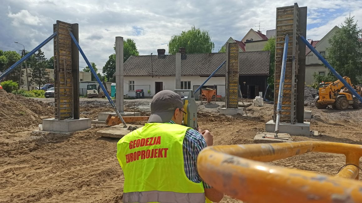 Pracownik w żółtej kamizelce z napisem 'Geodezja Europrojekt' obsługuje tachimetr na placu budowy, w tle widoczne szalunki kolumn i budynek w trakcie budowy.