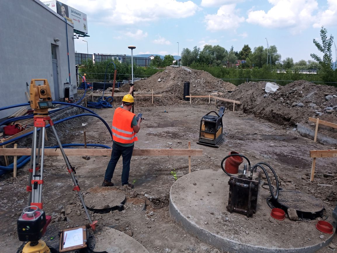 Pracownik w pomarańczowej kamizelce i żółtym kasku używa smartfona na placu budowy, w tle geodezyjny tachimetr na statywie, wykop fundamentowy z drewnianym szalunkiem, ubijarka, hałda ziemi i budynek.