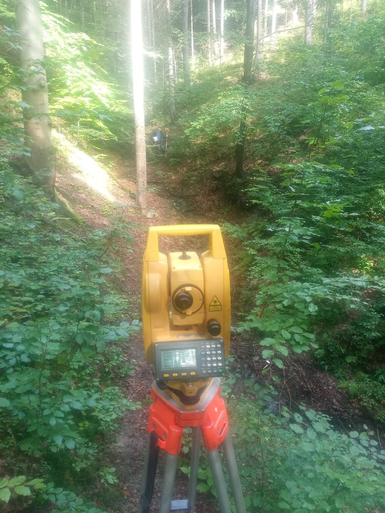 Żółty teodolit na czerwonym statywie w gęstym, zielonym lesie, pomiar w terenie z zaroślami i drzewami w tle.