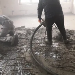 Dwóch pracowników rozprowadza masę na podłodze w pomieszczeniu, jeden używa węża do wylewania, drugi wyrównuje szpachlą, widoczna siatka zbrojeniowa i folia pod wylewką.