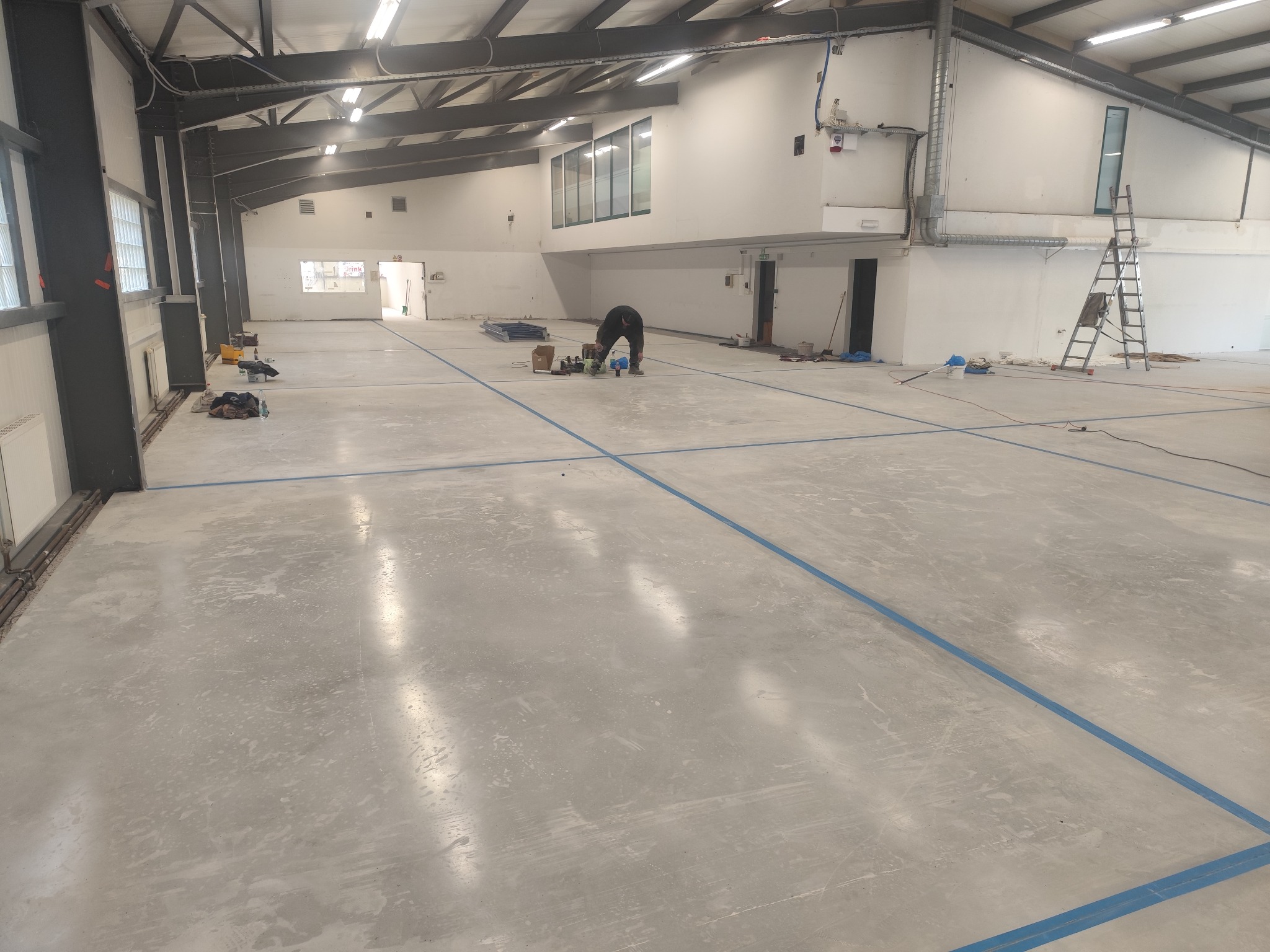 Pracownik przygotowuje posadzkę w hali sportowej z wyznaczonymi liniami boiska. Widoczne narzędzia i materiały budowlane. Hala oświetlona światłem dziennym i sztucznym.