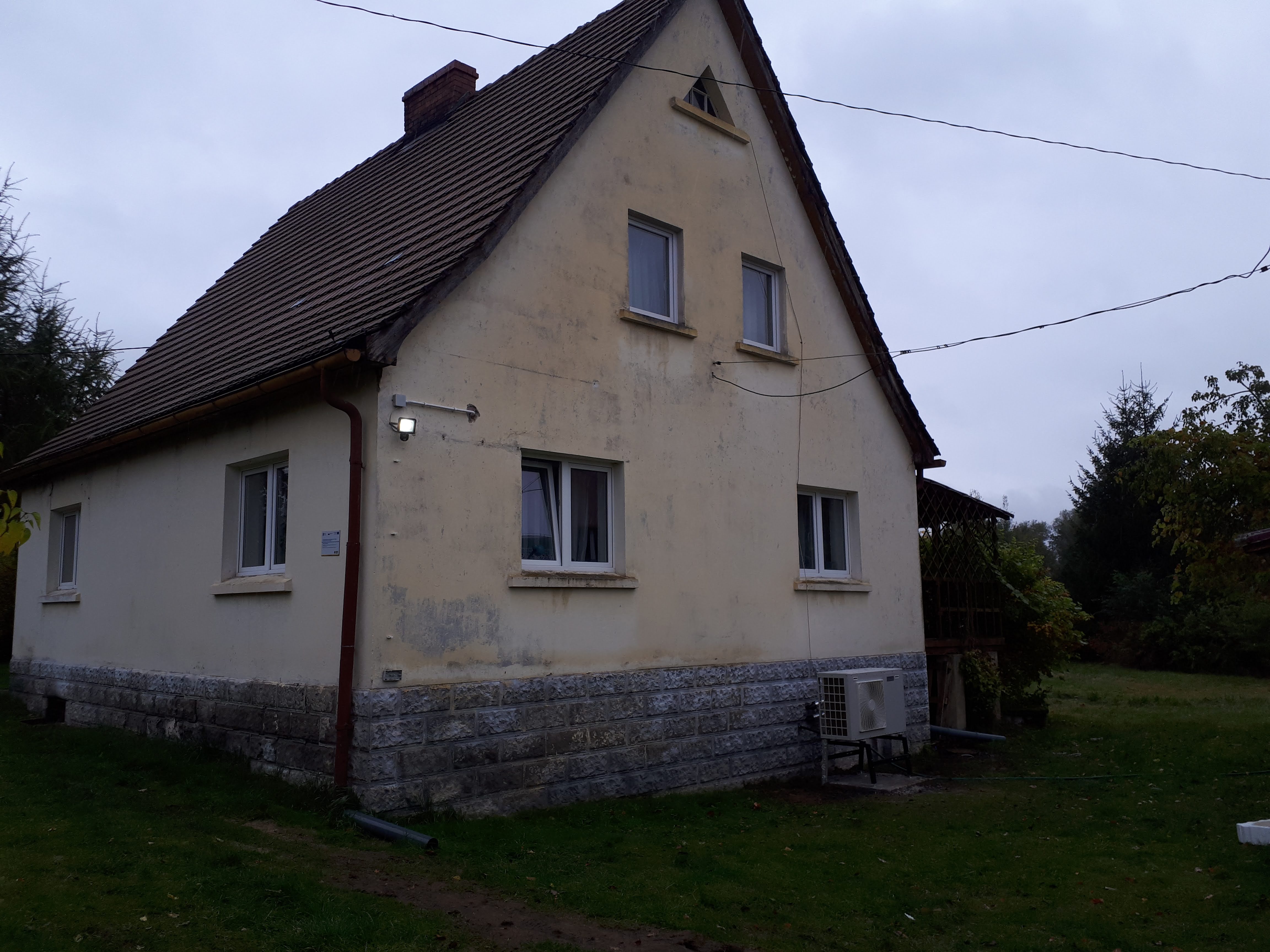 Dom jednorodzinny z zamontowaną jednostką zewnętrzną pompy ciepła na metalowym stelażu, widoczną przy ścianie budynku z ceglaną podmurówką.