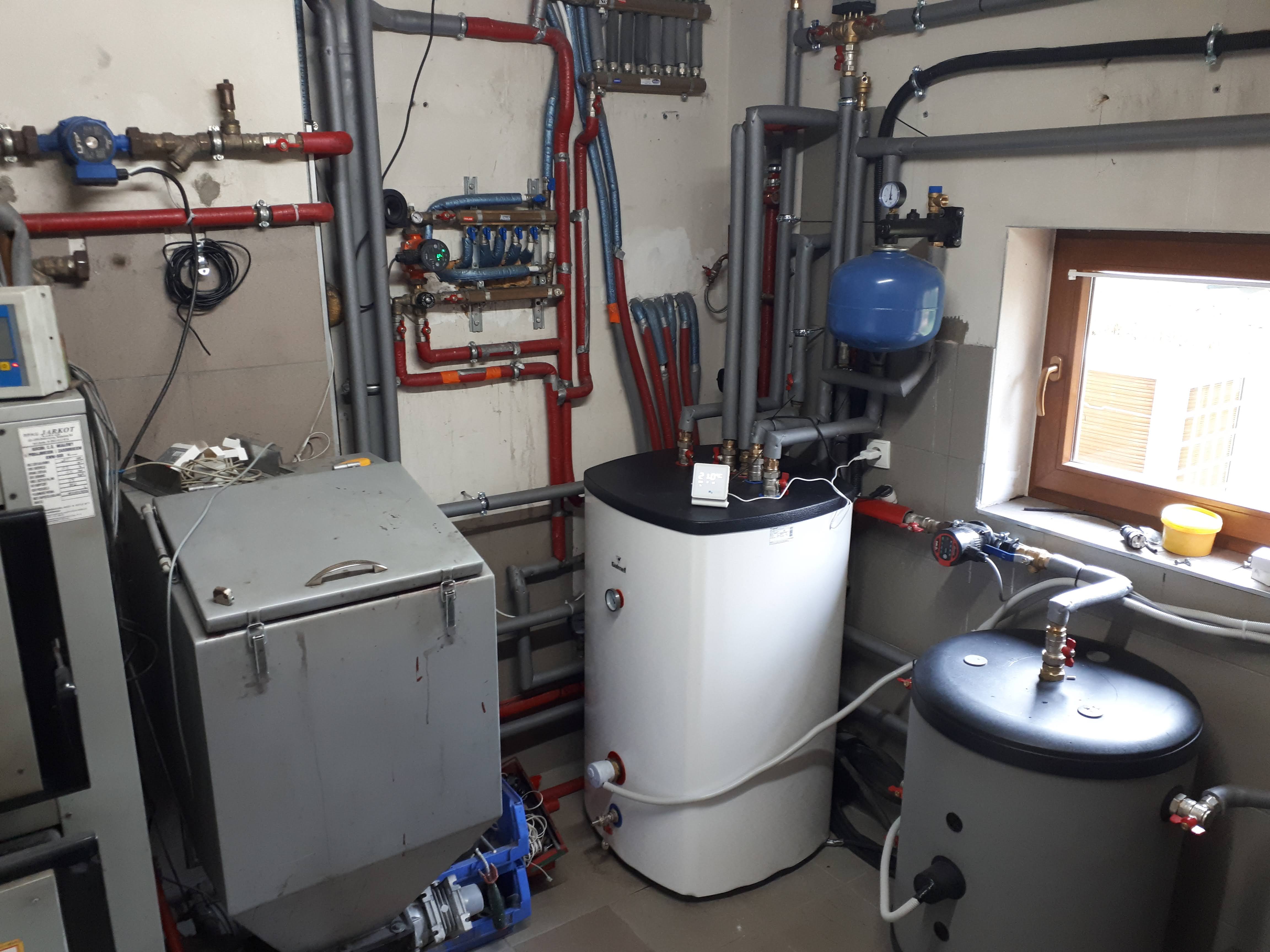 Kotłownia z widocznym białym zbiornikiem buforowym z czarnym blatem, szarym zasobnikiem ciepłej wody, licznymi rurami w kolorach szarym i czerwonym oraz rozdzielaczem podłogówki na ścianie.