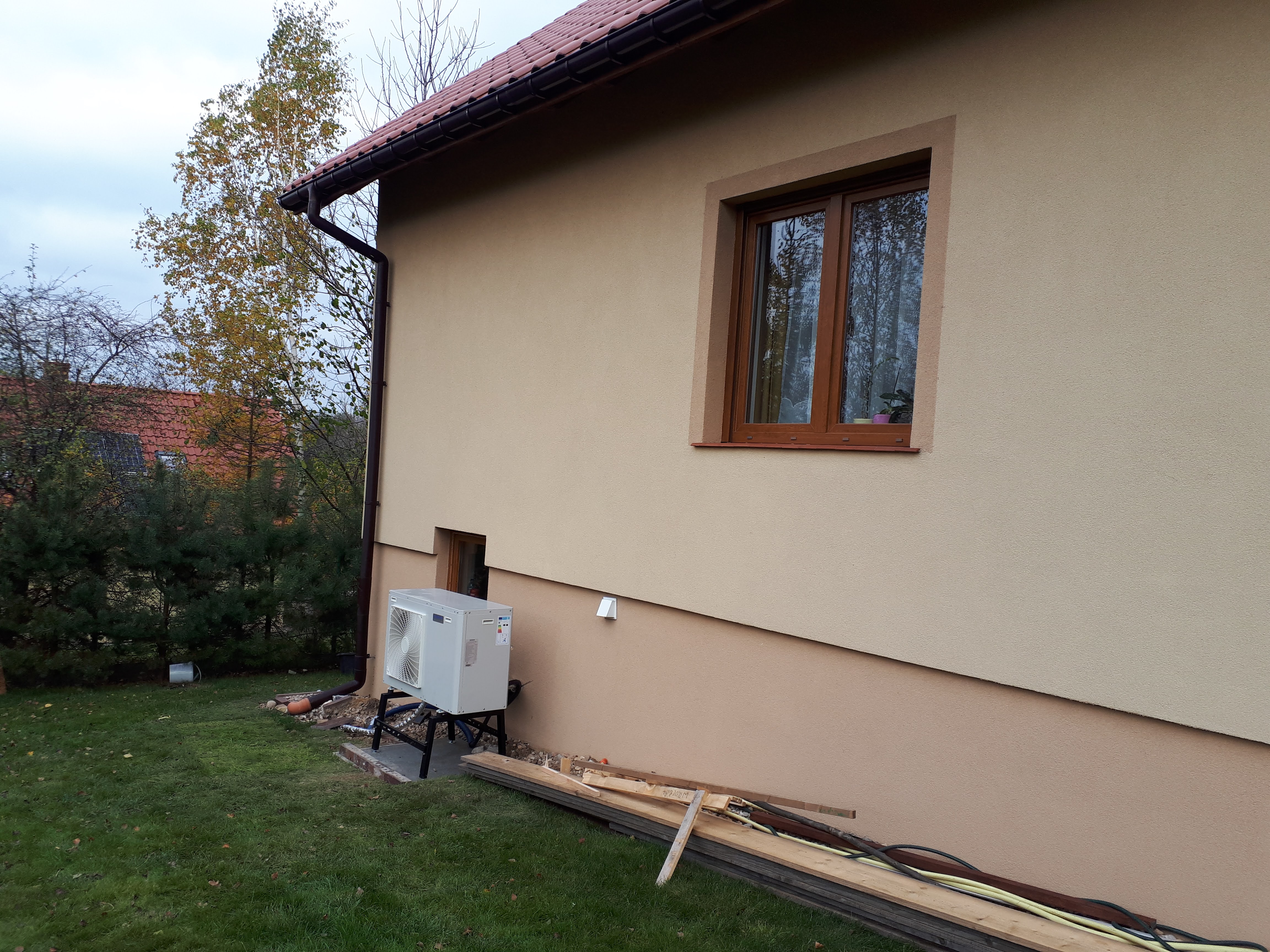 Zewnętrzna jednostka pompy ciepła na metalowej konstrukcji, umieszczona przy ścianie domu z beżową elewacją, widoczne deski i przewody na trawniku.