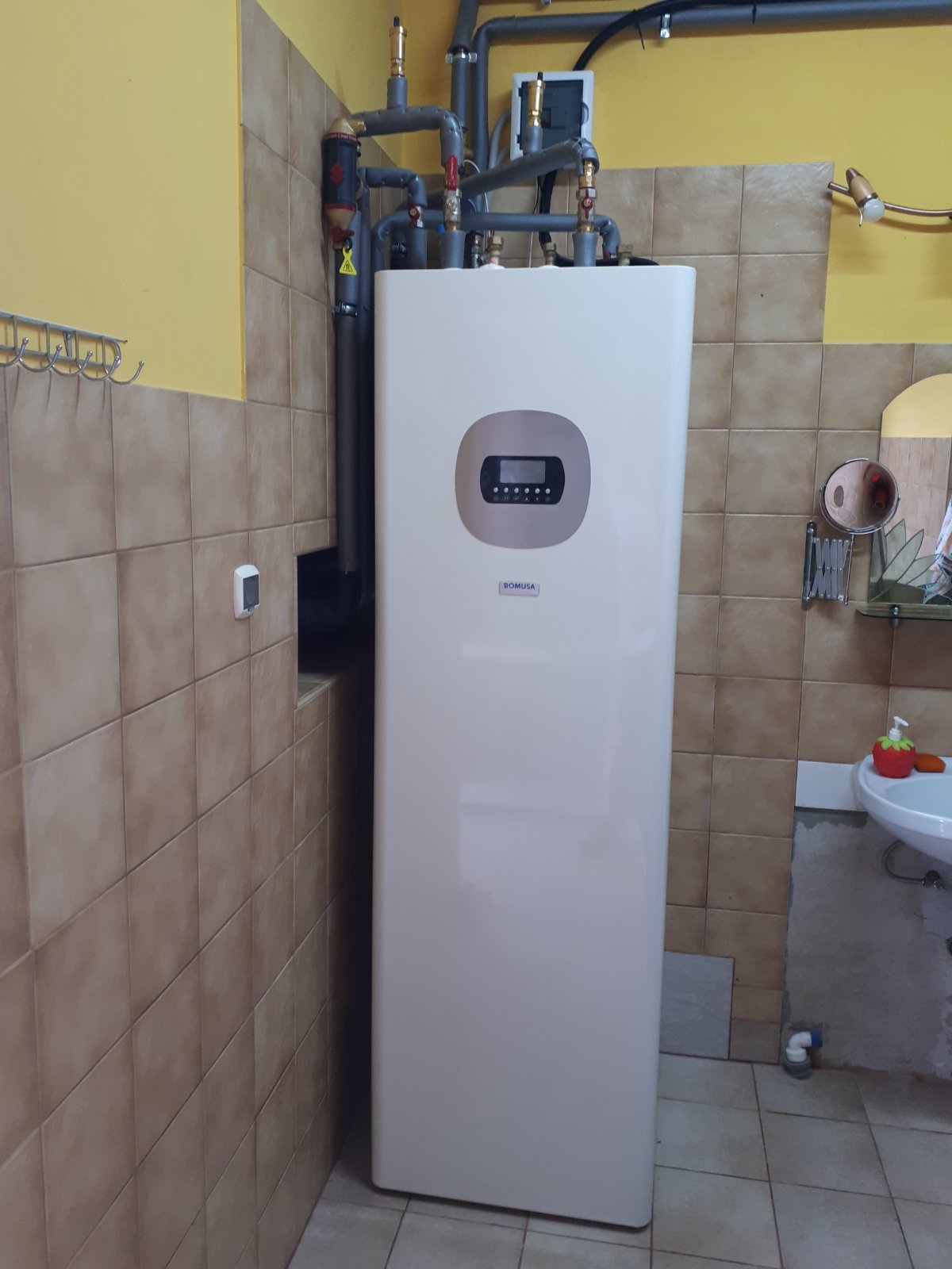 Biała, pionowa jednostka z szarym panelem sterowania i logo Domusa, podłączona do rur, zamontowana w pomieszczeniu z beżowymi płytkami i żółtą ścianą, obok umywalki.
