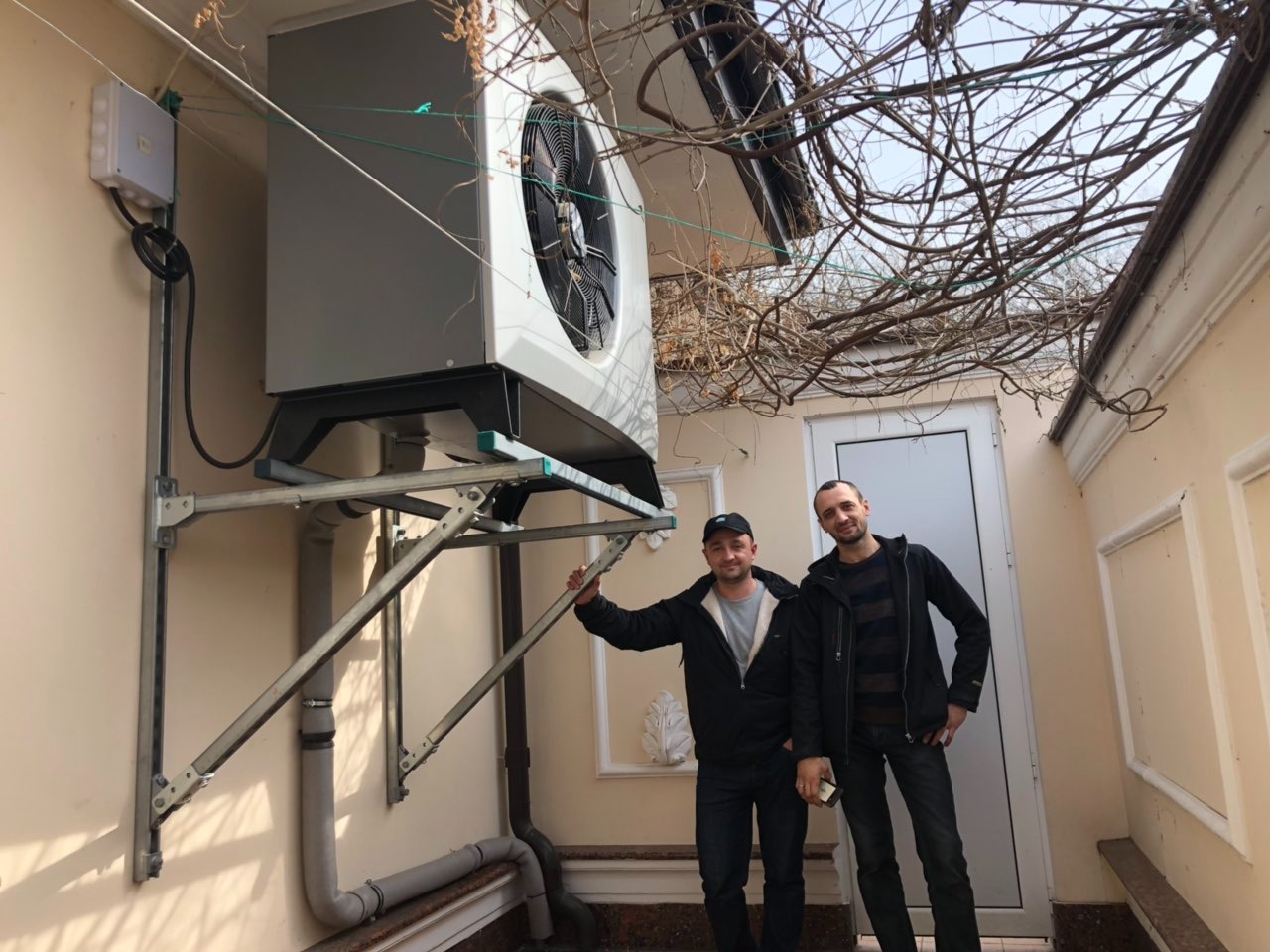 Zewnętrzna jednostka z wentylatorem zamontowana na wspornikach na ścianie budynku, widoczni dwaj mężczyźni stojący pod urządzeniem, nad nimi gałęzie drzewa.