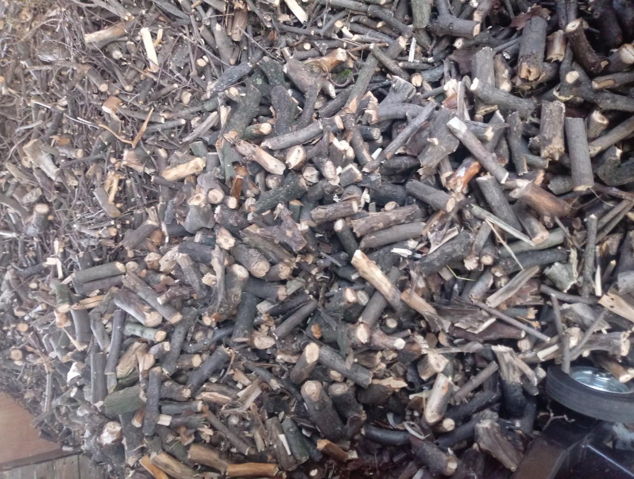 Stos pociętych gałęzi i kawałków drewna, widoczne koło wózka transportowego w prawym dolnym rogu.