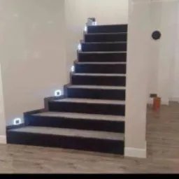 Schody wyłożone ciemną wykładziną z podświetleniem LED na każdym stopniu, widok z dołu, jasne ściany i podłoga imitująca drewno.