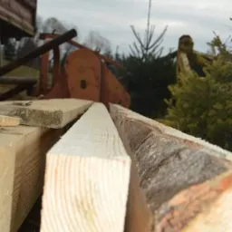 Świeżo przetarte deski z widoczną korą na jednej z nich, ułożone obok siebie, w tle maszyna do obróbki drewna i drzewa.