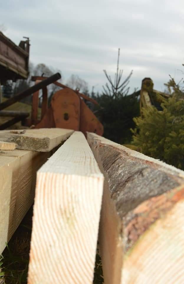 Świeżo przetarte deski z widoczną korą na jednej z nich, ułożone obok siebie, w tle maszyna do obróbki drewna i drzewa.