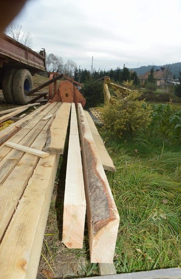 Świeżo przetarte deski leżące na przyczepie, z widoczną korą na krawędziach, w tle zieleń i zabudowania.
