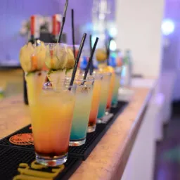 Rząd kolorowych drinków z owocowymi dekoracjami i czarnymi słomkami na barze, oświetlonych fioletowym światłem.