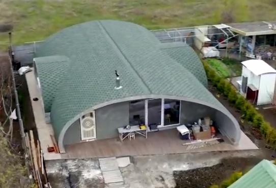 Nietypowy dom kopułowy z zielonym dachem z gontu bitumicznego, widok z lotu ptaka, w tle zielone pole i zabudowania gospodarcze.