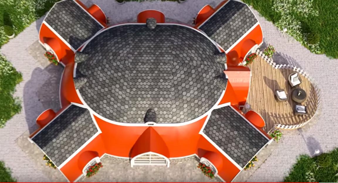 Wizualizacja z góry domu o nietypowym, okrągłym kształcie z czerwonymi ścianami i szarym dachem z dachówki, z przylegającym drewnianym tarasem z meblami ogrodowymi.