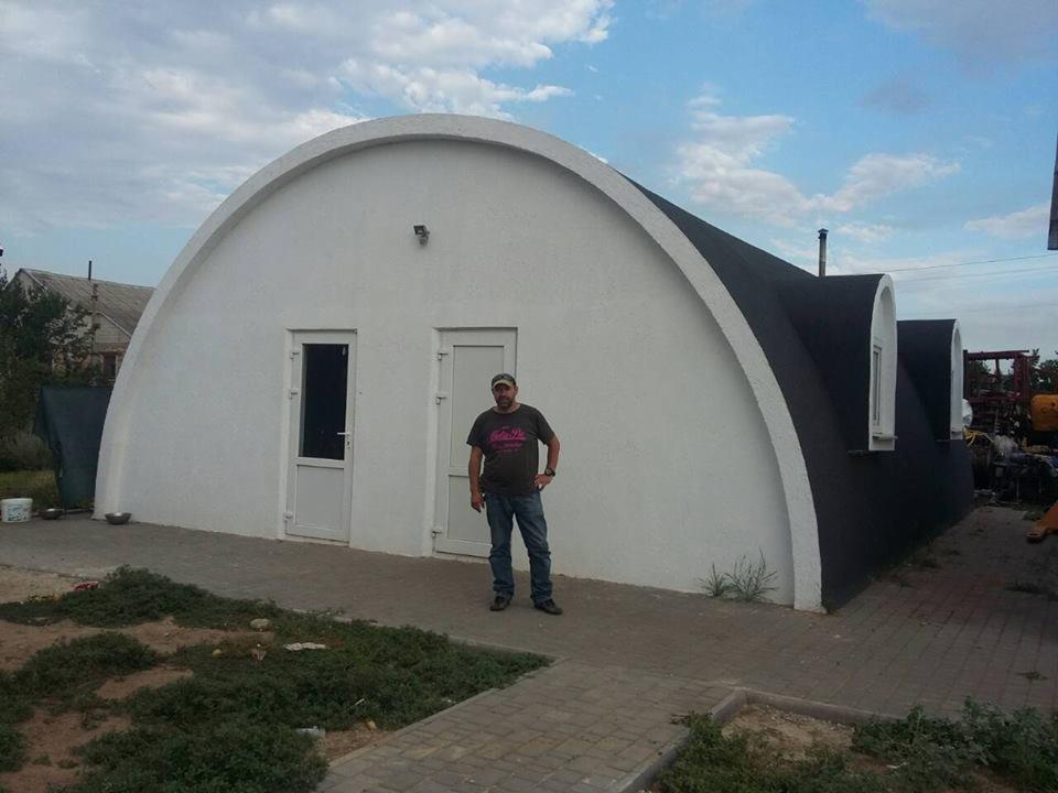 Nietypowy dom modułowy o łukowej konstrukcji, z dwoma wejściami i osobą stojącą przed budynkiem, widoczne elementy wykończeniowe i fragment placu budowy w tle.