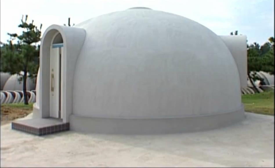 Betonowy dom kopułowy z wejściem łukowym i ciemną wycieraczką, otoczony zielenią i innymi kopułami w tle.