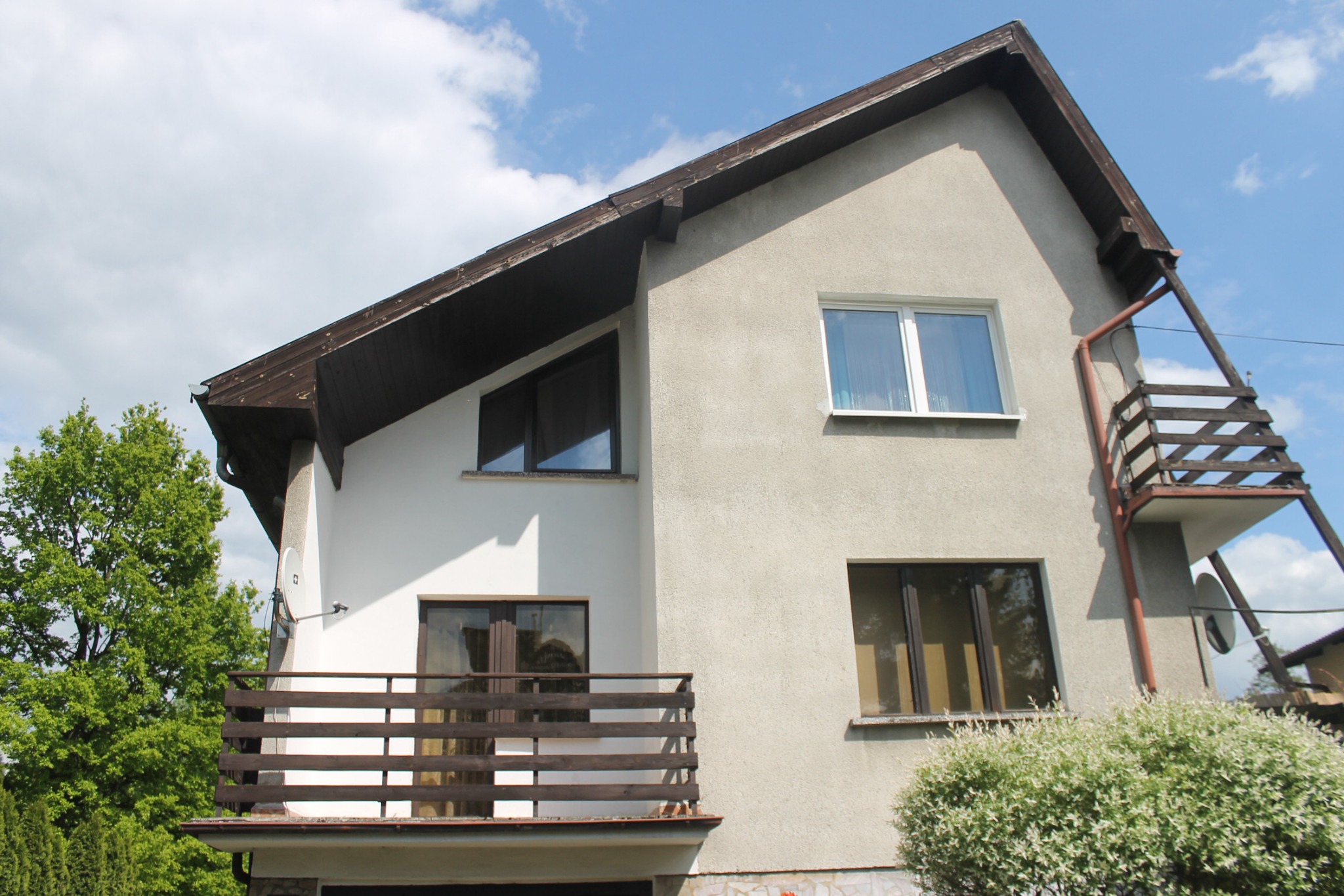 Dwukondygnacyjny dom z jasną elewacją, drewnianą balustradą balkonu i zewnętrznymi schodami ewakuacyjnymi z metalową rynną, widziany z dołu na tle błękitnego nieba.