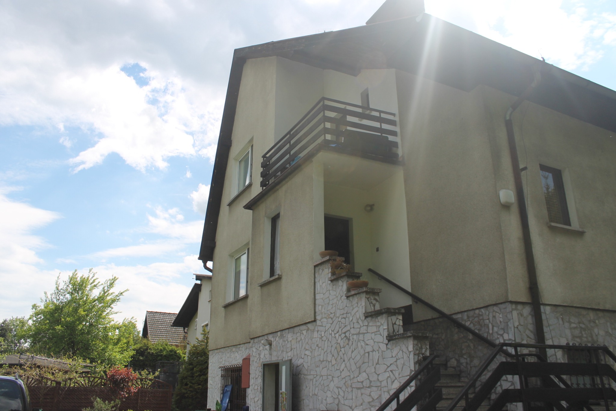 Dwupiętrowy dom z balkonem i schodami wejściowymi wykończonymi kamieniem dekoracyjnym, widok z poziomu gruntu na tle błękitnego nieba z chmurami.