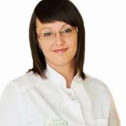 Uśmiechnięta dietetyczka w okularach i białym fartuchu, z identyfikatorem z logo, na jasnym tle. Portret w neutralnym oświetleniu.