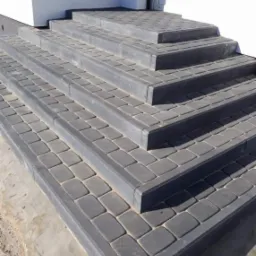 Schody wejściowe wyłożone grafitową kostką brukową, z widocznym świeżym cementem na obrzeżach, prowadzące do białego budynku z zielonymi roletami.