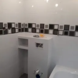 Biała łazienka z mozaikową listwą dekoracyjną, zabudowa geberit z półką, widoczne przyłącza wodne i fragment wanny.