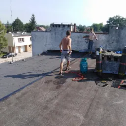 Dwóch dekarzy bez koszulek na płaskim dachu z papy, butla gazowa i zrolowane pasy papy w tle, widok na okoliczne budynki i drzewa w słoneczny dzień.