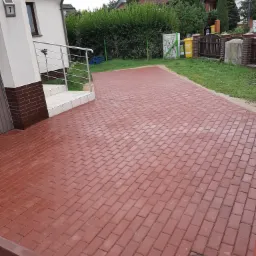 Czerwona kostka brukowa ułożona na podjeździe przed domem z numerem 7, widoczny właz kanalizacyjny i fragment schodów wejściowych.