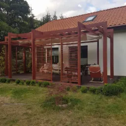 Drewniana konstrukcja z lamelami i zadaszeniem materiałowym przylegająca do domu z czerwoną dach&oacute;wką, otoczona zielenią.