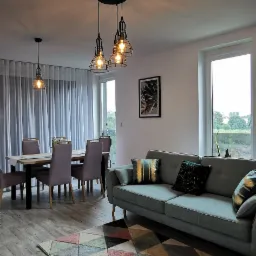 Jasne wnętrze salonu z drewnianą podłogą, szarą sofą z poduszkami dekoracyjnymi, stołem z krzesłami i dywanem w geometryczne wzory, oświetlone wiszącymi lampami z metalowymi kloszami.