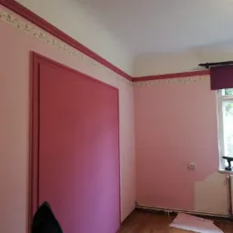 Pokój po odświeżeniu: ściany pomalowane na jasny róż, z ciemnoróżowymi akcentami przy suficie i wokół drzwi. Na parapecie w oknie stoi wiertarko-wkrętarka. Widoczny fragment grzejnika i podłogi.