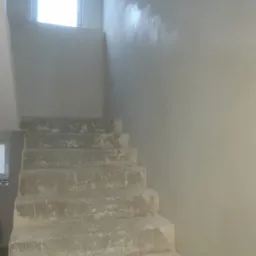 Schody w trakcie prac wykończeniowych, ściana częściowo pokryta warstwą wyrównującą, widoczne niedoskonałości na stopniach i ścianie, okno w tle.