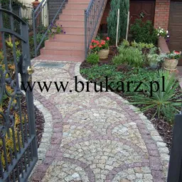 Kaskadowy chodnik z kostki granitowej w odcieniach szarości i różu, prowadzący od ozdobnej, otwartej bramy z kutego żelaza w kierunku schodów i garażu, otoczony rabatami z zielenią ozdobną...