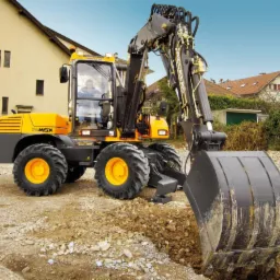 Żółto-szara koparko-ładowarka Mecalac 11cxi z operatorem w kabinie, podczas wykonywania wykopu w ziemi na tle zabudowy mieszkalnej.