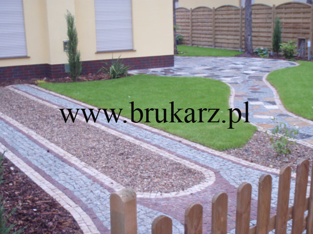 Ogród z alejkami wyłożonymi kostką granitową i żwirem, obrzeża z czerwonej kostki, trawnik, drewniany płotek, budynek z ceglaną podmurówką i roletami w tle.
