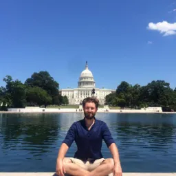 Młody mężczyzna siedzi w pozycji lotosu na tle budynku Kapitolu w Waszyngtonie, odbijającego się w wodzie.