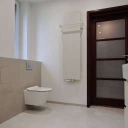 WB-Qube Waldemar Bednarczyk - Jasna łazienka po remoncie: podwieszana toaleta, beżowe płytki, grzejnik drabinkowy i ciemne drzwi z przeszkleniami. Minimalistyczny design, czyste linie.