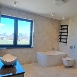 WB-Qube Waldemar Bednarczyk - Jasna łazienka z beżowymi płytkami imitującymi kamień, wolnostojąca wanna, umywalka nablatowa i grzejnik drabinkowy. Widok na okno z zielenią w tle.